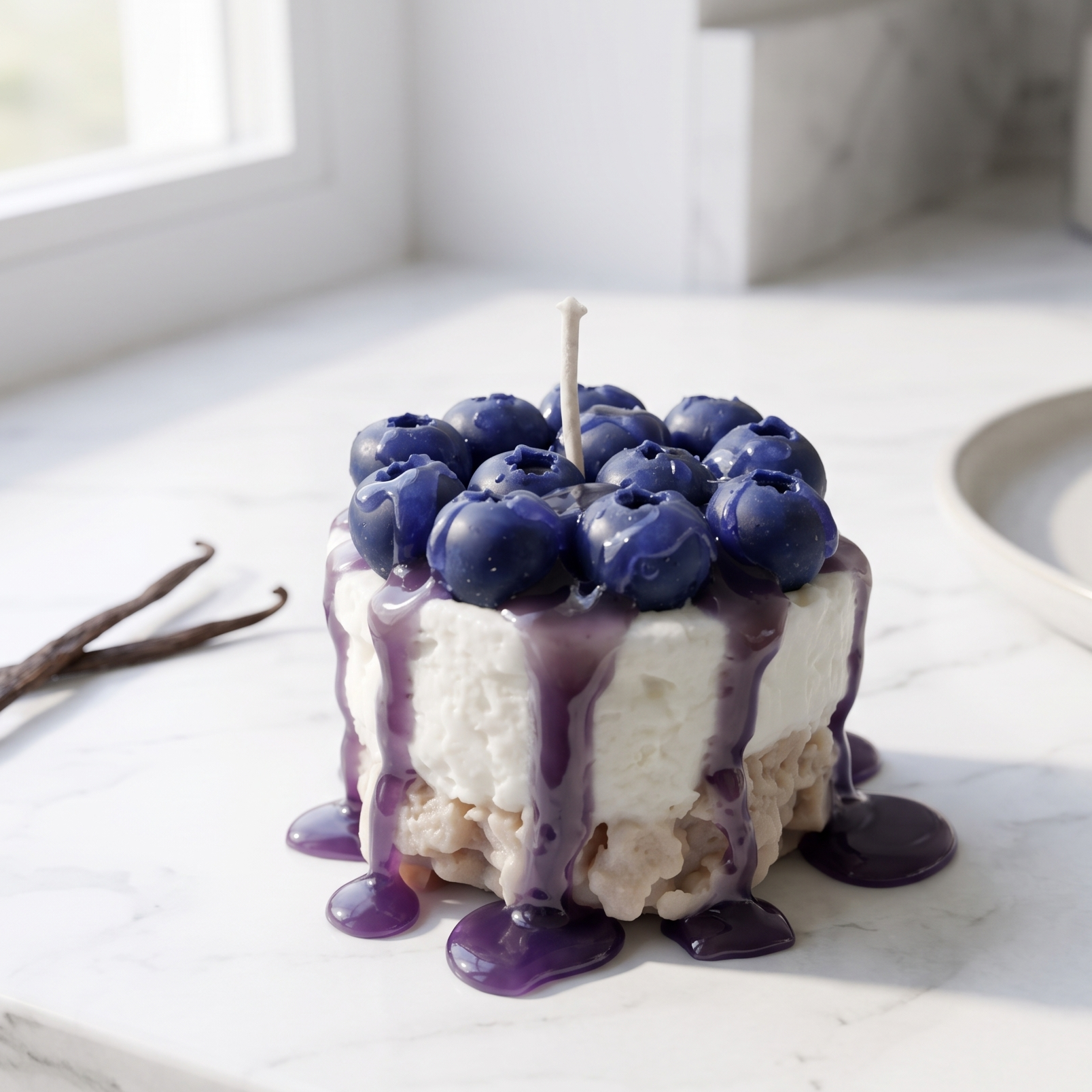 handcrafted Mini Blueberry Cheesecake Candle by SORA SCENT standing on a minimalist wooden coffee table, featuring a textured crumb base, smooth white cream layer, dripping purple blueberry glaze, and realistic deep-blue wax blueberries on top, next to a blurred lifestyle magazine.