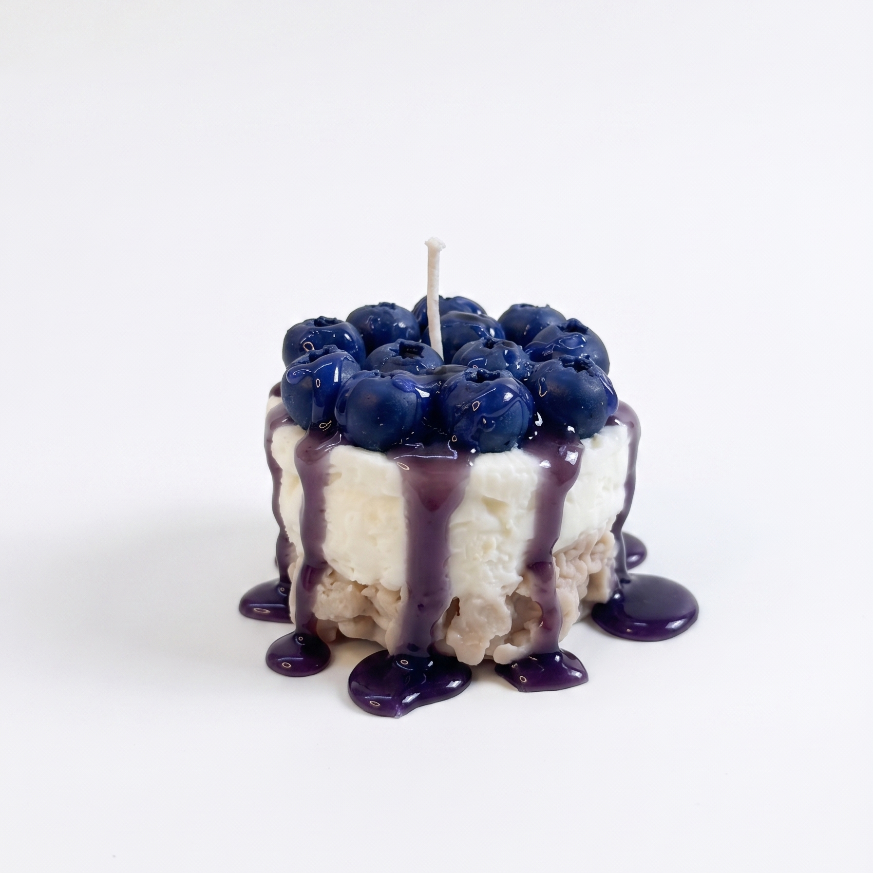 handcrafted Mini Blueberry Cheesecake Candle by SORA SCENT standing on a minimalist wooden coffee table, featuring a textured crumb base, smooth white cream layer, dripping purple blueberry glaze, and realistic deep-blue wax blueberries on top, next to a blurred lifestyle magazine.