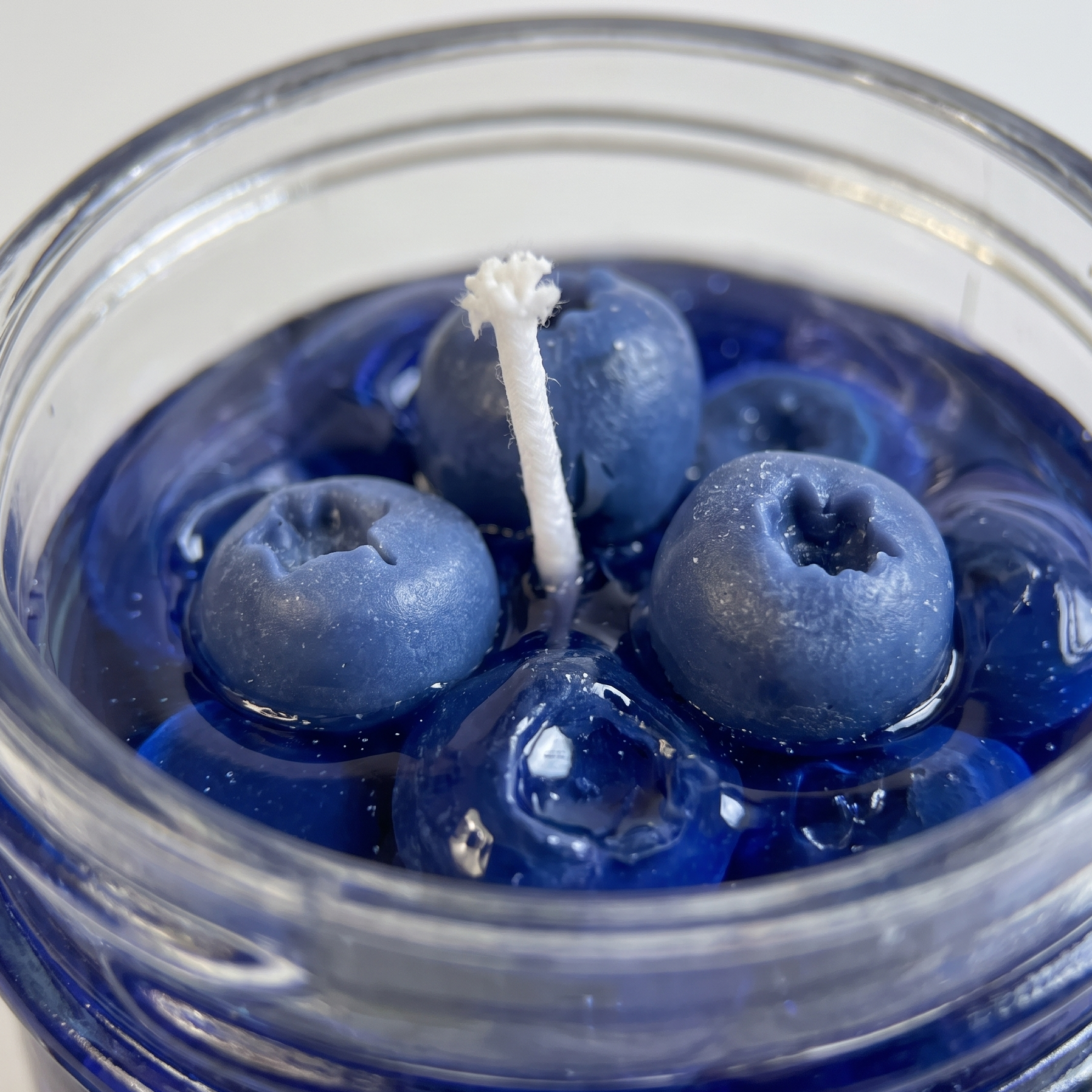 The sora-scent-blueberry-gel-jelly-candle-white-background.jpg.png presents a handcrafted Wild Blueberry Preserves Candle by SORA SCENT in a clear glass jar on a white background, filled with translucent blue jelly wax and realistic wax blueberries, surrounded by loose decorative blueberries.