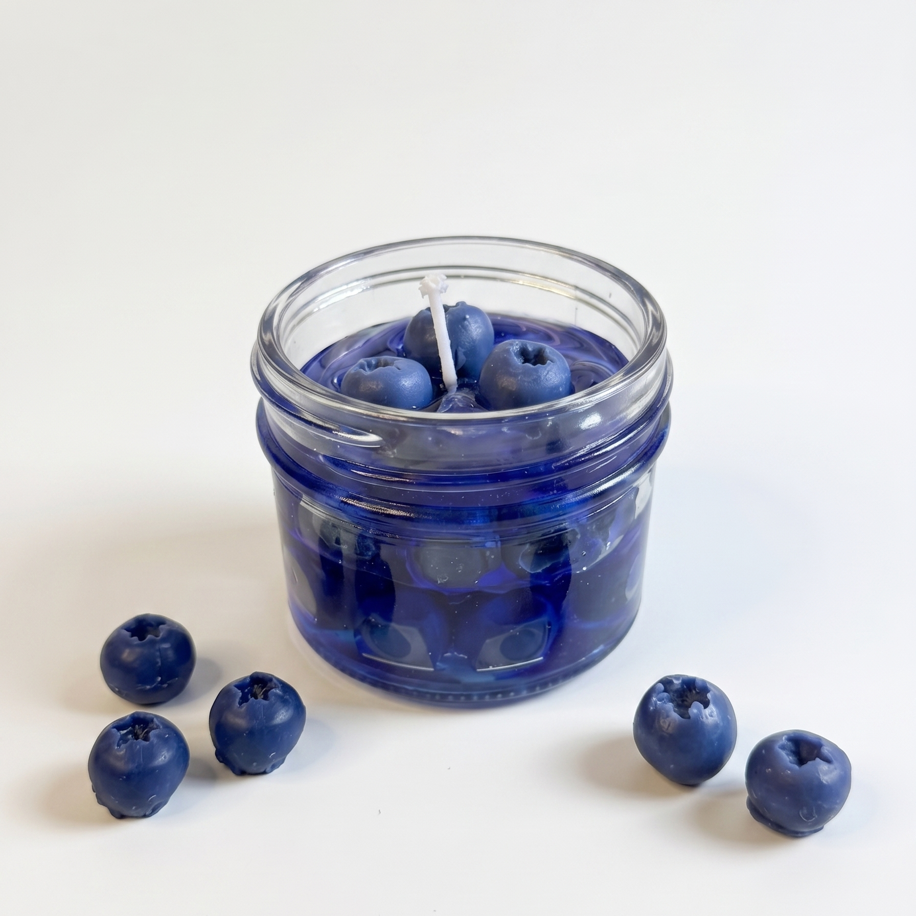 The sora-scent-blueberry-gel-jelly-candle-white-background.jpg.png presents a handcrafted Wild Blueberry Preserves Candle by SORA SCENT in a clear glass jar on a white background, filled with translucent blue jelly wax and realistic wax blueberries, surrounded by loose decorative blueberries.
