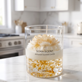 shows a handcrafted Golden Peony Lavender Luxury Glass Candle by SORA SCENT sitting on a white marble kitchen countertop, featuring gold leaf gel wax, a creamy soy base, and a sculpted white wax flower.