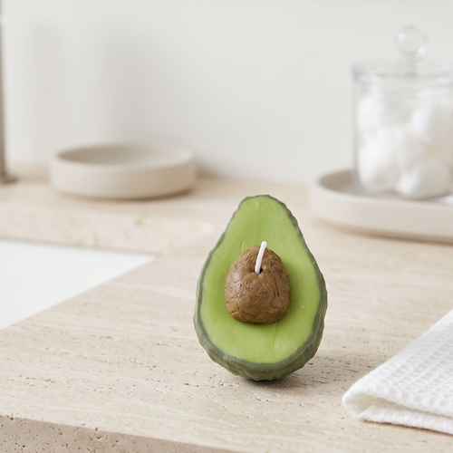 The sora-scent-half-cut-avocado-shaped-candle-bathroom-vanity.jpg.png displays a hyper-realistic, handcrafted Half-Cut Hass Avocado Candle by SORA SCENT sitting on a beige travertine bathroom counter, featuring a textured green rind and a realistic brown pit, next to a white cotton towel with a blurred glass jar of cotton balls in the background.