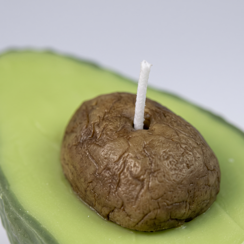 The sora-scent-half-cut-avocado-shaped-candle-bathroom-vanity.jpg.png displays a hyper-realistic, handcrafted Half-Cut Hass Avocado Candle by SORA SCENT sitting on a beige travertine bathroom counter, featuring a textured green rind and a realistic brown pit, next to a white cotton towel with a blurred glass jar of cotton balls in the background.