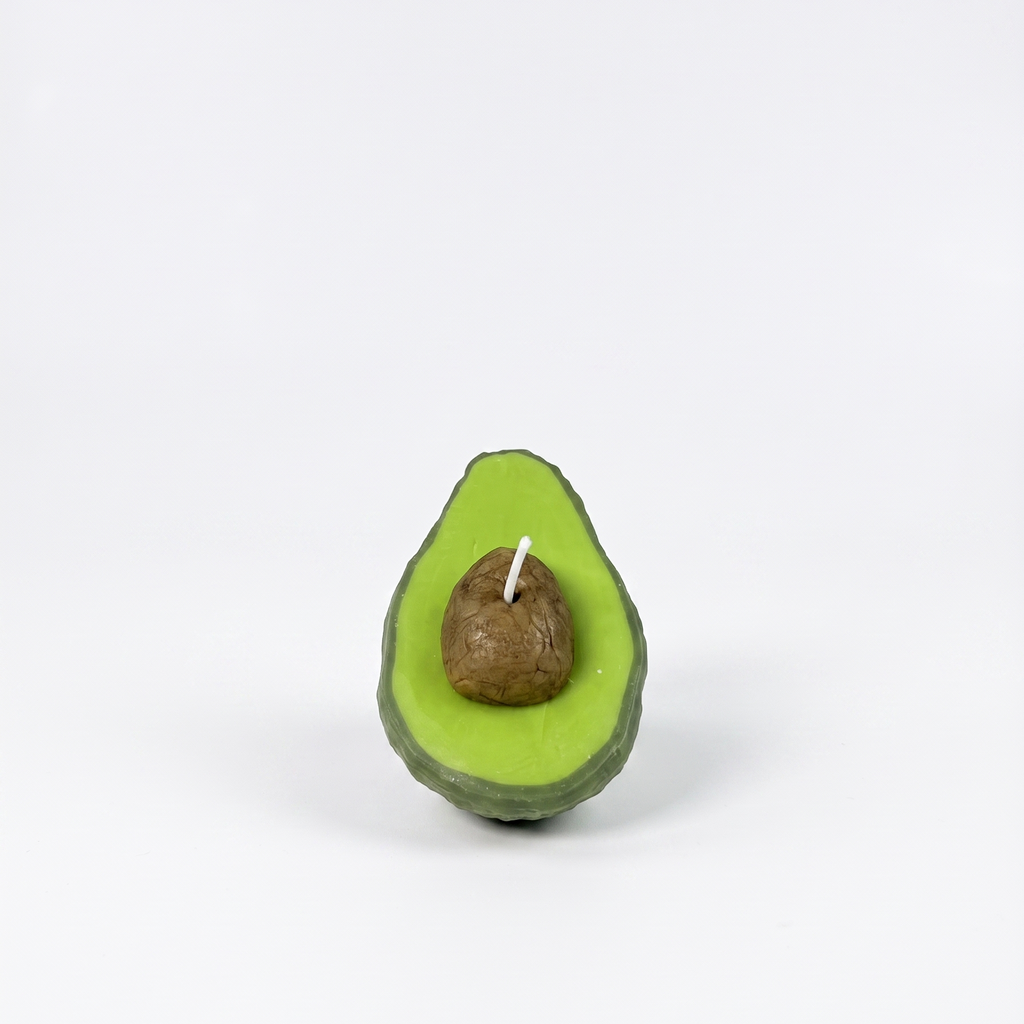 The sora-scent-half-cut-avocado-shaped-candle-bathroom-vanity.jpg.png displays a hyper-realistic, handcrafted Half-Cut Hass Avocado Candle by SORA SCENT sitting on a beige travertine bathroom counter, featuring a textured green rind and a realistic brown pit, next to a white cotton towel with a blurred glass jar of cotton balls in the background.