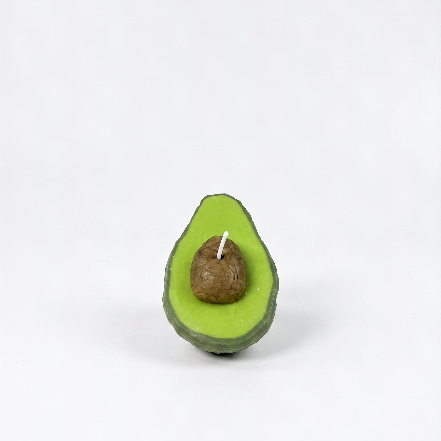 The sora-scent-half-cut-avocado-shaped-candle-bathroom-vanity.jpg.png displays a hyper-realistic, handcrafted Half-Cut Hass Avocado Candle by SORA SCENT sitting on a beige travertine bathroom counter, featuring a textured green rind and a realistic brown pit, next to a white cotton towel with a blurred glass jar of cotton balls in the background.