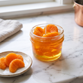 shows a handcrafted Sweet Mandarin Marmalade Candle by SORA SCENT in a glass jar on a marble kitchen island, filled with golden translucent jelly wax and realistic wax mandarin segments, next to a small ceramic plate with more loose wax orange slices.