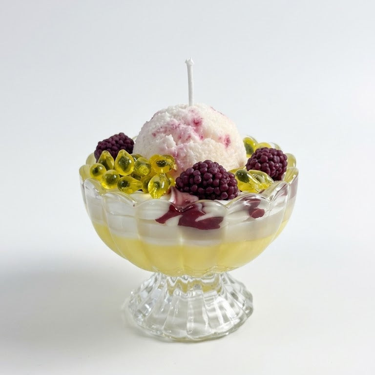 shows a handcrafted Passion Mulberry Ice Cream Sundae Candle by SORA SCENT in a clear glass goblet on a white sunlit windowsill, featuring layered yellow and white wax, realistic whipped cream, a pink ice cream scoop, glossy gel-wax passion fruit seeds, and dark purple wax mulberries.