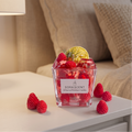 shows a handcrafted Strawberry Fizz Cocktail Candle by SORA SCENT sitting on a white wooden bedside nightstand in a cozy bedroom, featuring realistic wax strawberries and a lemon garnish, with loose decorative berries resting nearby.