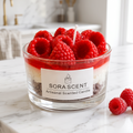 showcases a lit handcrafted Raspberry Mousse Tart Candle by SORA SCENT on a white wooden nightstand in a cozy, softly blurred bedroom setting, featuring distinct cookie crumb and cream layers topped with glowing red wax raspberries, with loose decorative wax berries resting beside it.