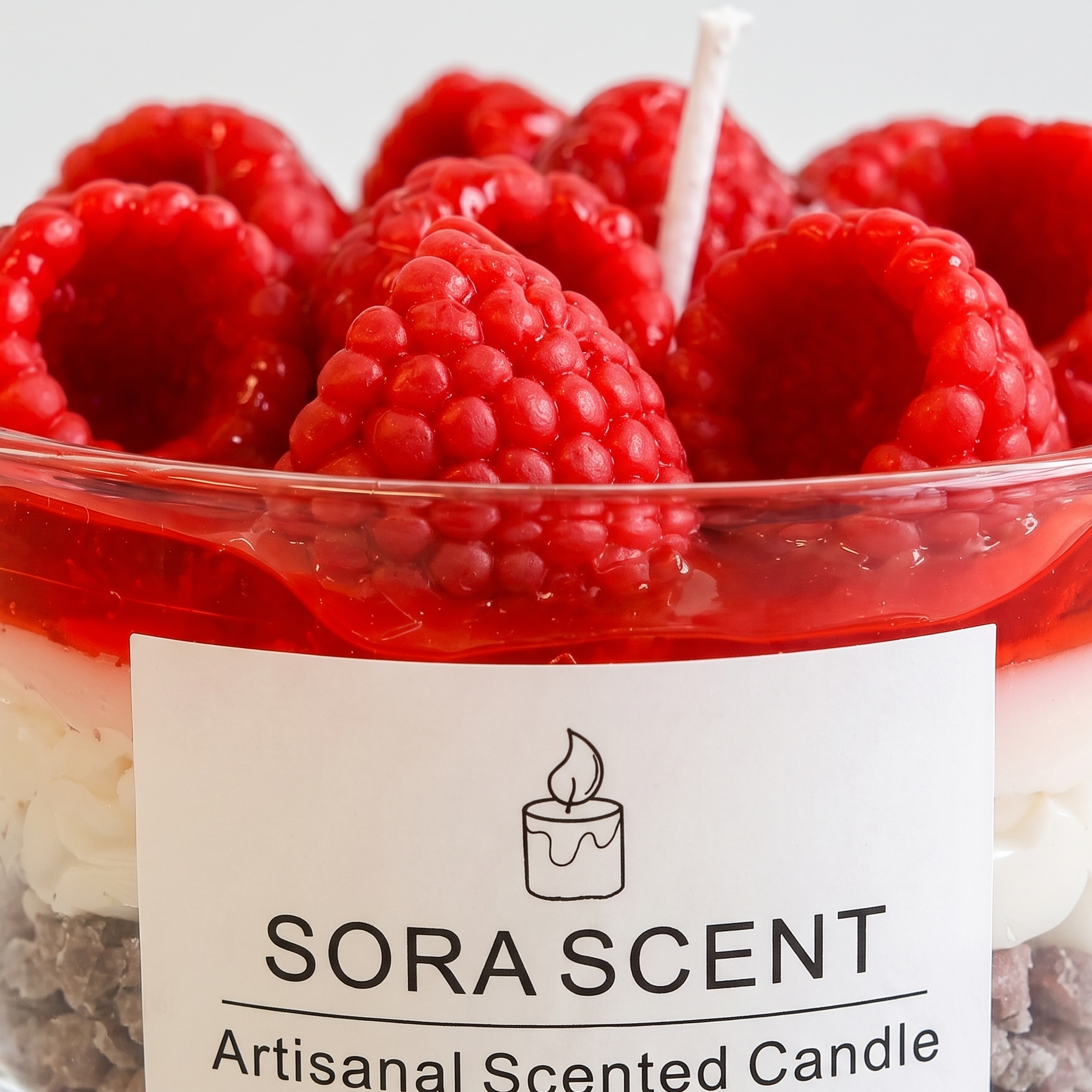 showcases a lit handcrafted Raspberry Mousse Tart Candle by SORA SCENT on a white wooden nightstand in a cozy, softly blurred bedroom setting, featuring distinct cookie crumb and cream layers topped with glowing red wax raspberries, with loose decorative wax berries resting beside it.