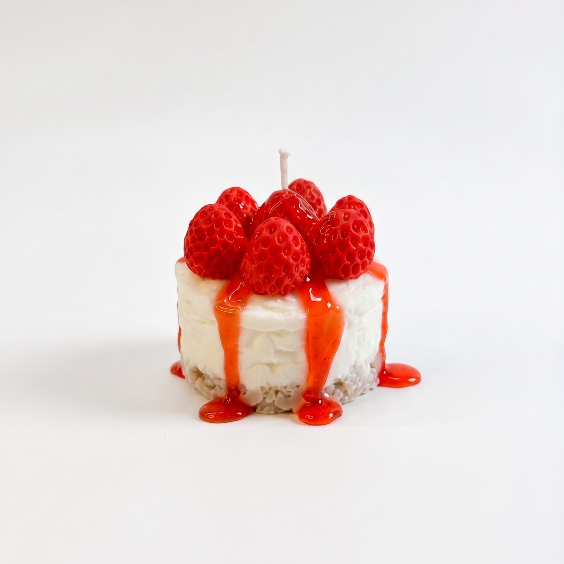 shows a hyper-realistic, handcrafted Mini Strawberry Cheesecake Candle by SORA SCENT standing on a minimalist wooden coffee table in soft sunlight, featuring a textured crumb crust base, a smooth white cream layer, dripping red strawberry glaze, and realistic red wax strawberries on top, with a blurred book in the background.