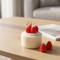 shows a lit handcrafted Mini Strawberry Mousse Cake Candle by SORA SCENT sitting on a ceramic dish on a wooden bedside nightstand, featuring creamy white soy wax, whipped cream, and realistic wax strawberries with loose berries beside it.